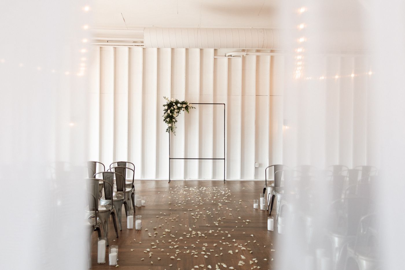 Indoor wedding ceremony setup at 14TENN in Nashville featuring a minimalist metal arch with greenery, white draped walls, scattered rose petals, and candlelit aisle.